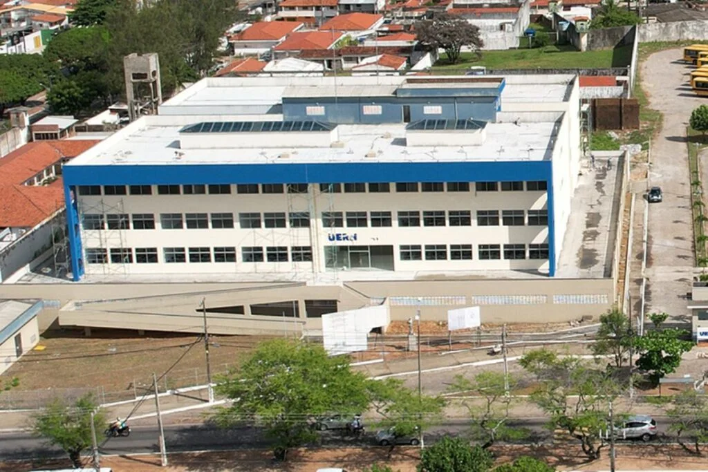Vista aérea do campus da Uern Natal, com prédio de fachada branca e detalhes azuis, cercado por ruas, árvores e imóveis residenciais