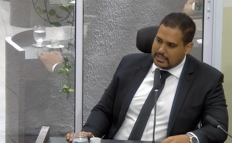 Michael Diniz, de terno escuro e gravata, sentado e falando ao microfone na Câmara de Parnamirim